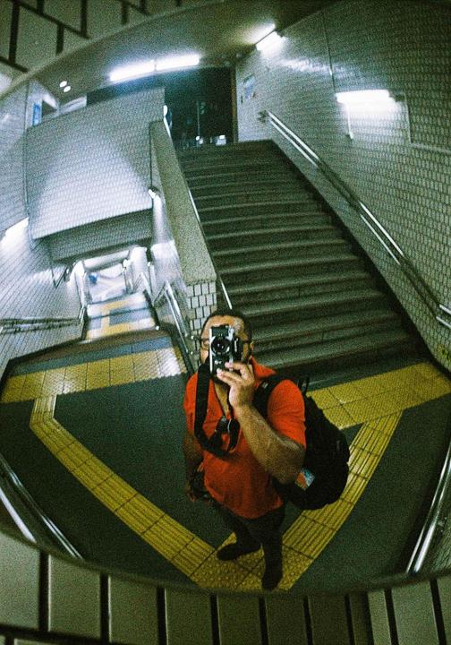 A man in a red shirt takes a self-portrait in a distorted reflection of a subway station.
