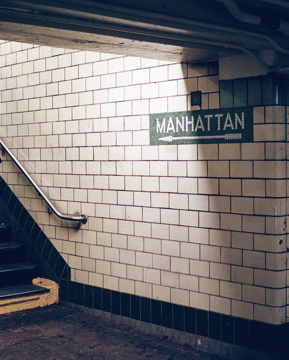A Manhattan subway sign with an arrow points to the right on a tiled wall next to a staircase.