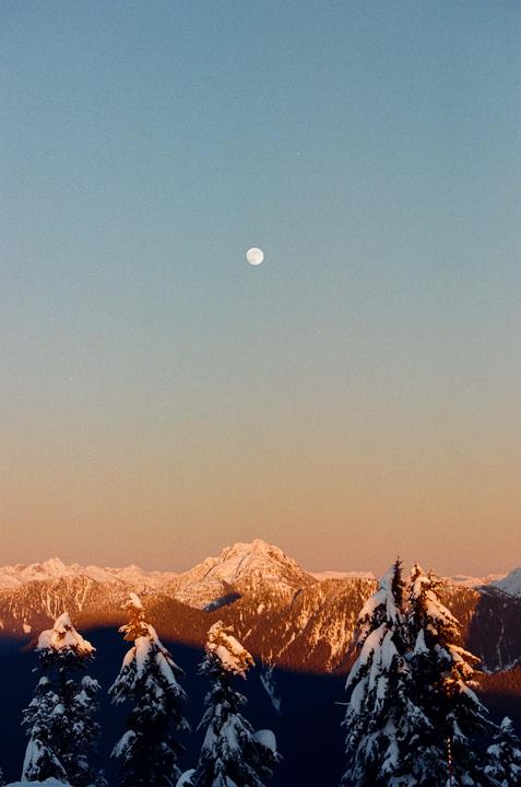 A full moon hangs in a clear sky above snow-covered mountains and pine trees.