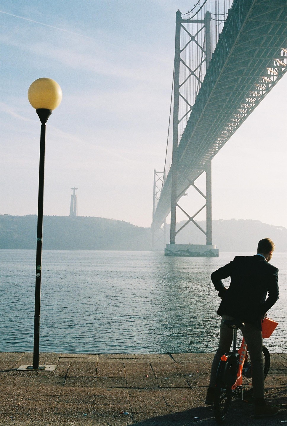 A man on a bicycle looks out at a suspension bridge over a body of water with a statue on a distant hill.
