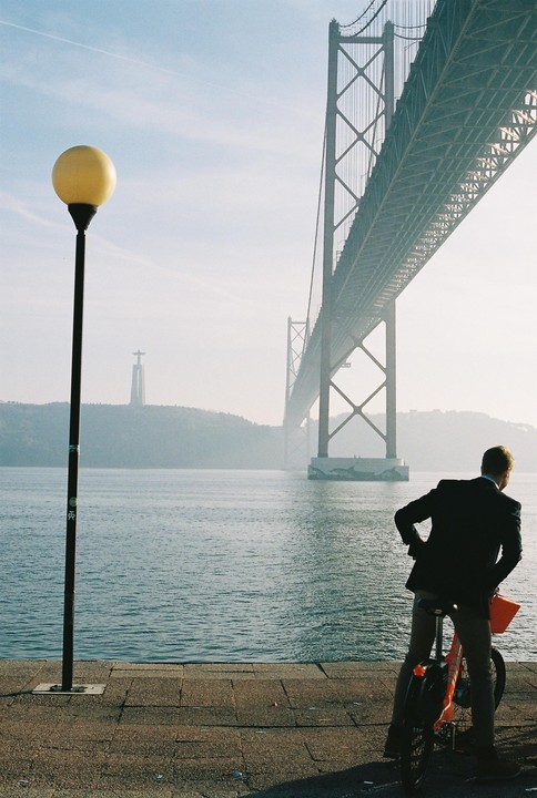 A man on a bicycle looks out at a suspension bridge over a body of water with a statue on a distant hill.