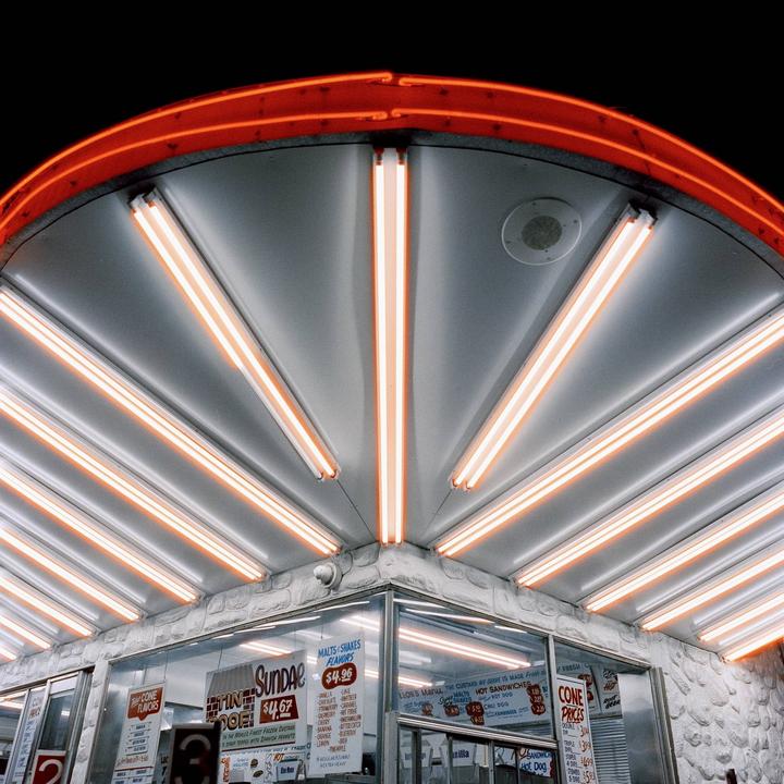 A brightly lit ice cream stand with neon lights and menu boards is shown at night.