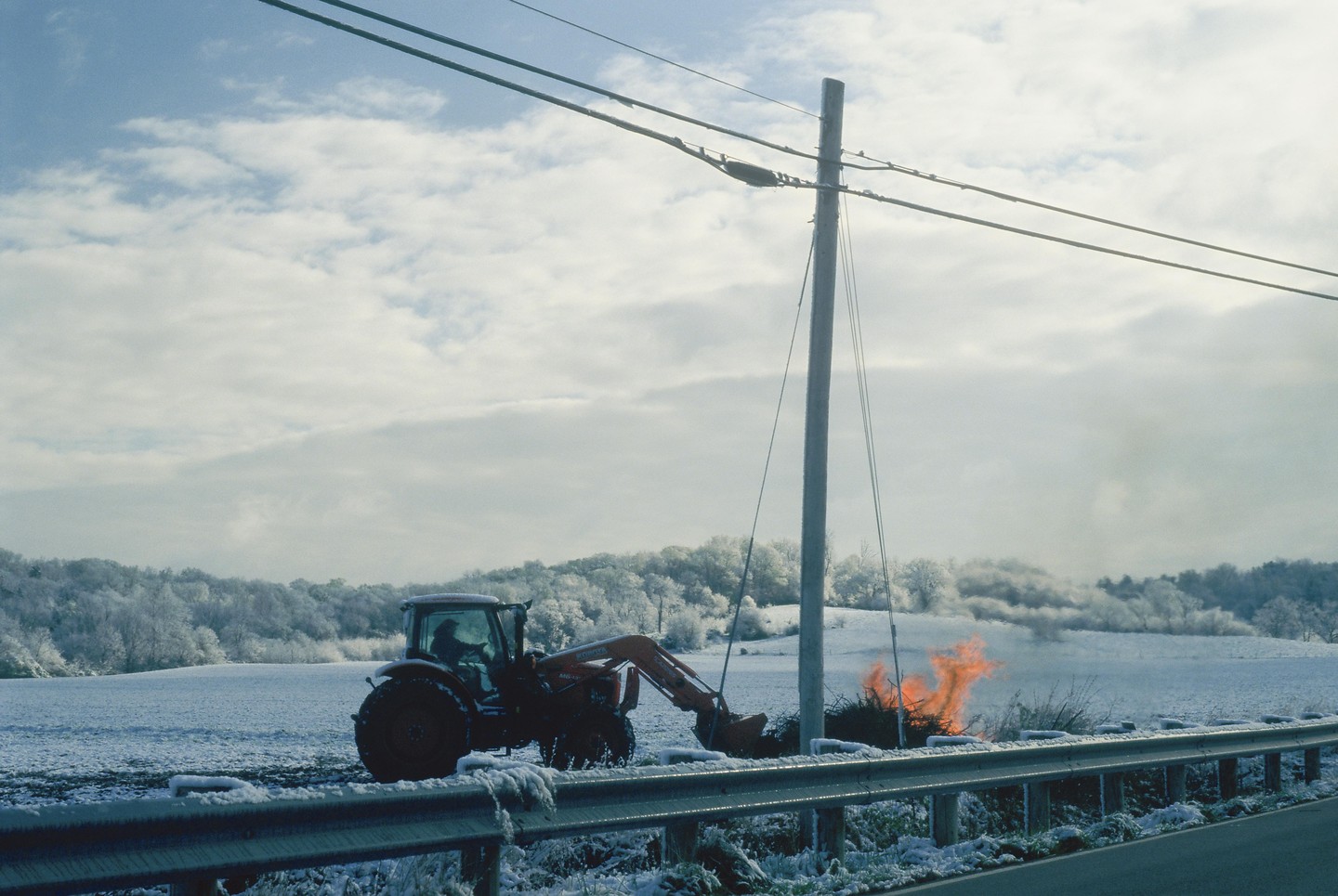 A tractor is parked next to a burning pile of brush in a snowy field with a forest in the background.
