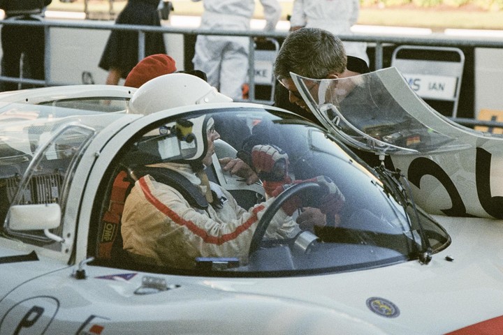 A man in a racing suit and helmet sits in a white race car while another man leans into the open cockpit.