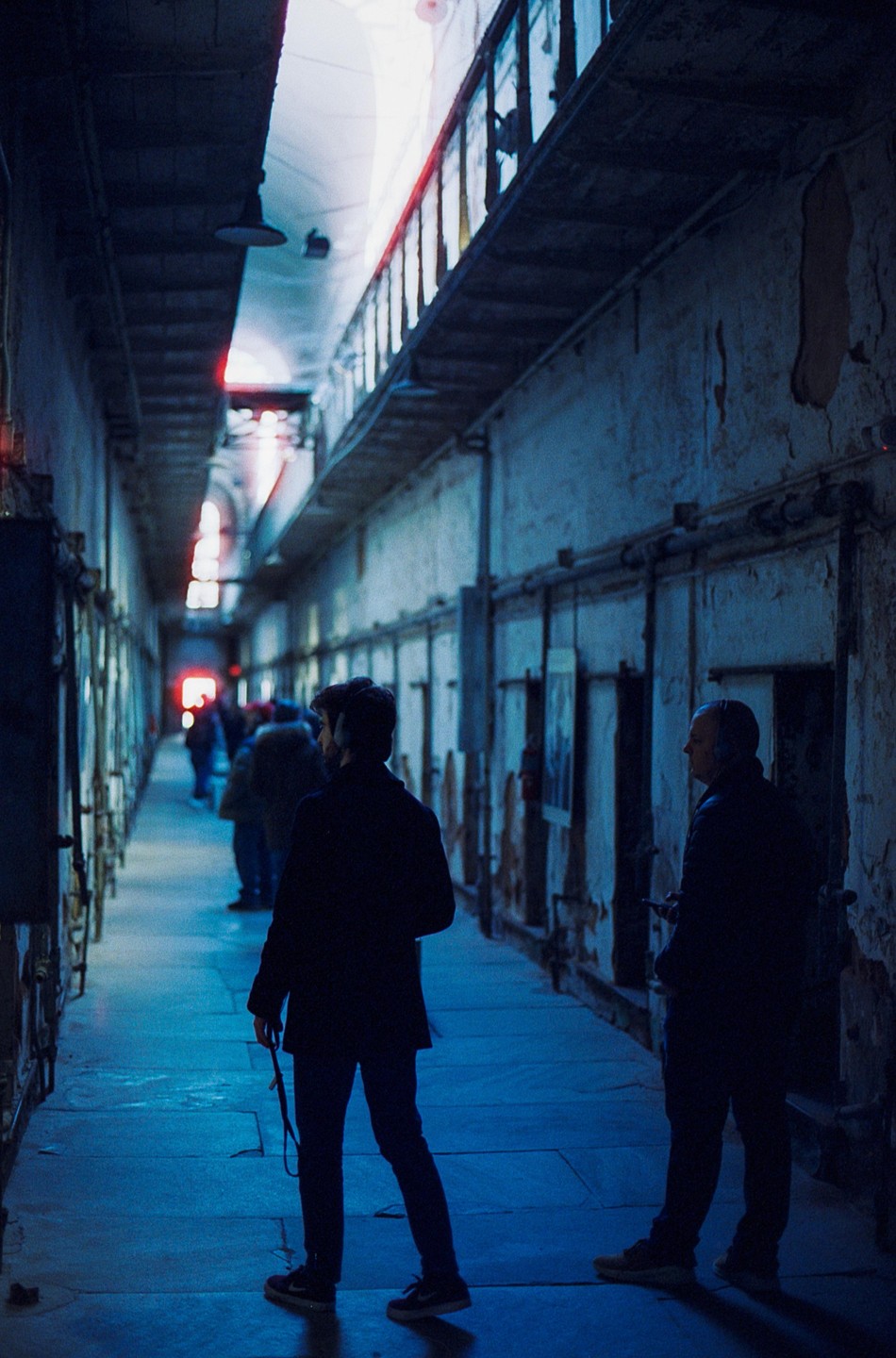 Two men stand in a long, narrow, blue-lit corridor with rows of cells on either side.