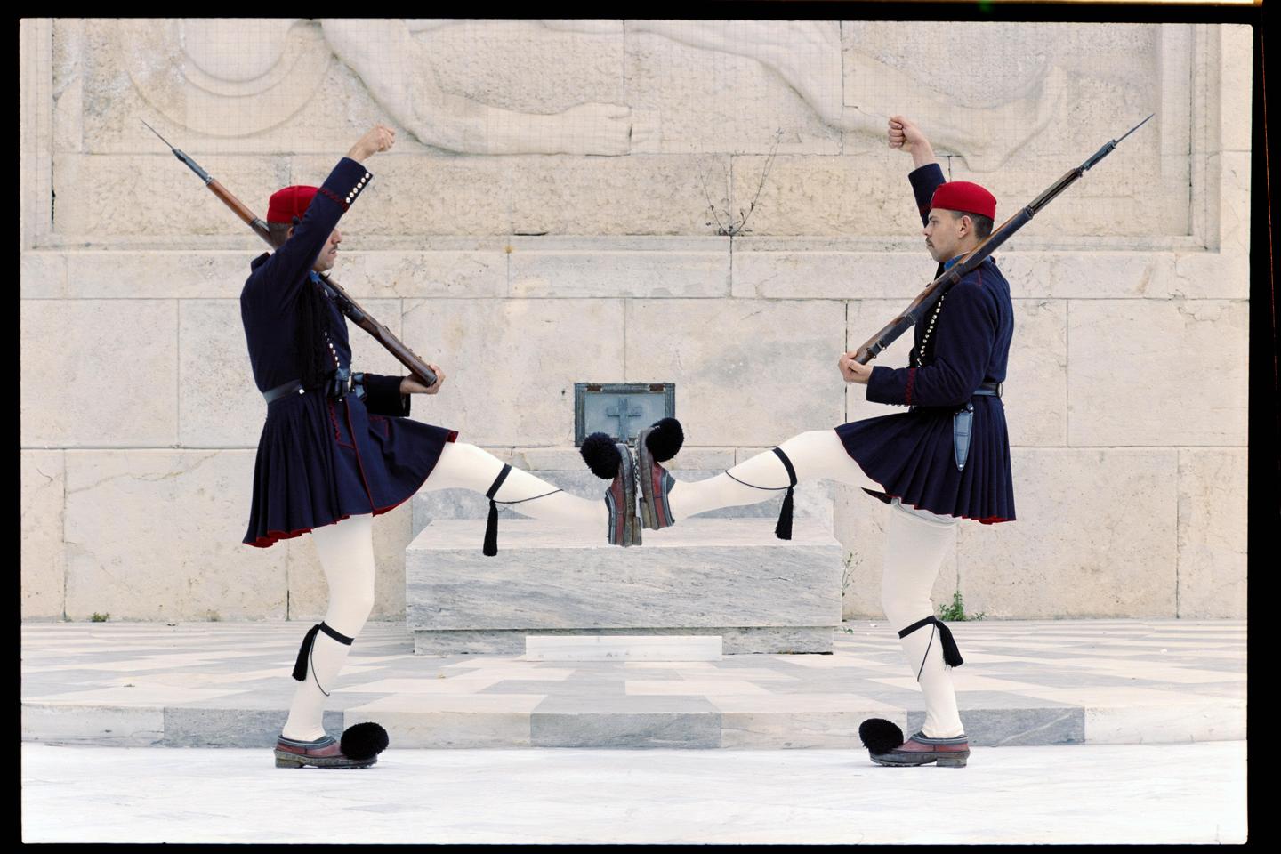 Two men in traditional uniform with rifles perform a synchronized high-kick in front of a stone wall.