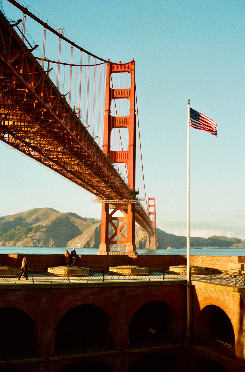The Golden Gate Bridge is visible from Fort Point, with an American flag flying in the foreground.