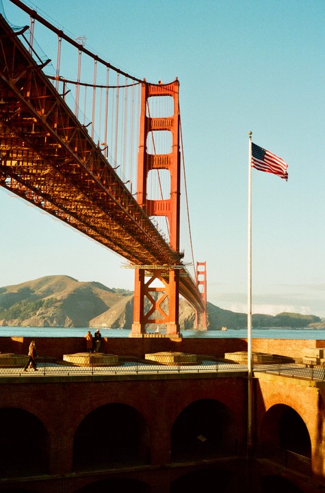 The Golden Gate Bridge is visible from Fort Point, with an American flag flying in the foreground.