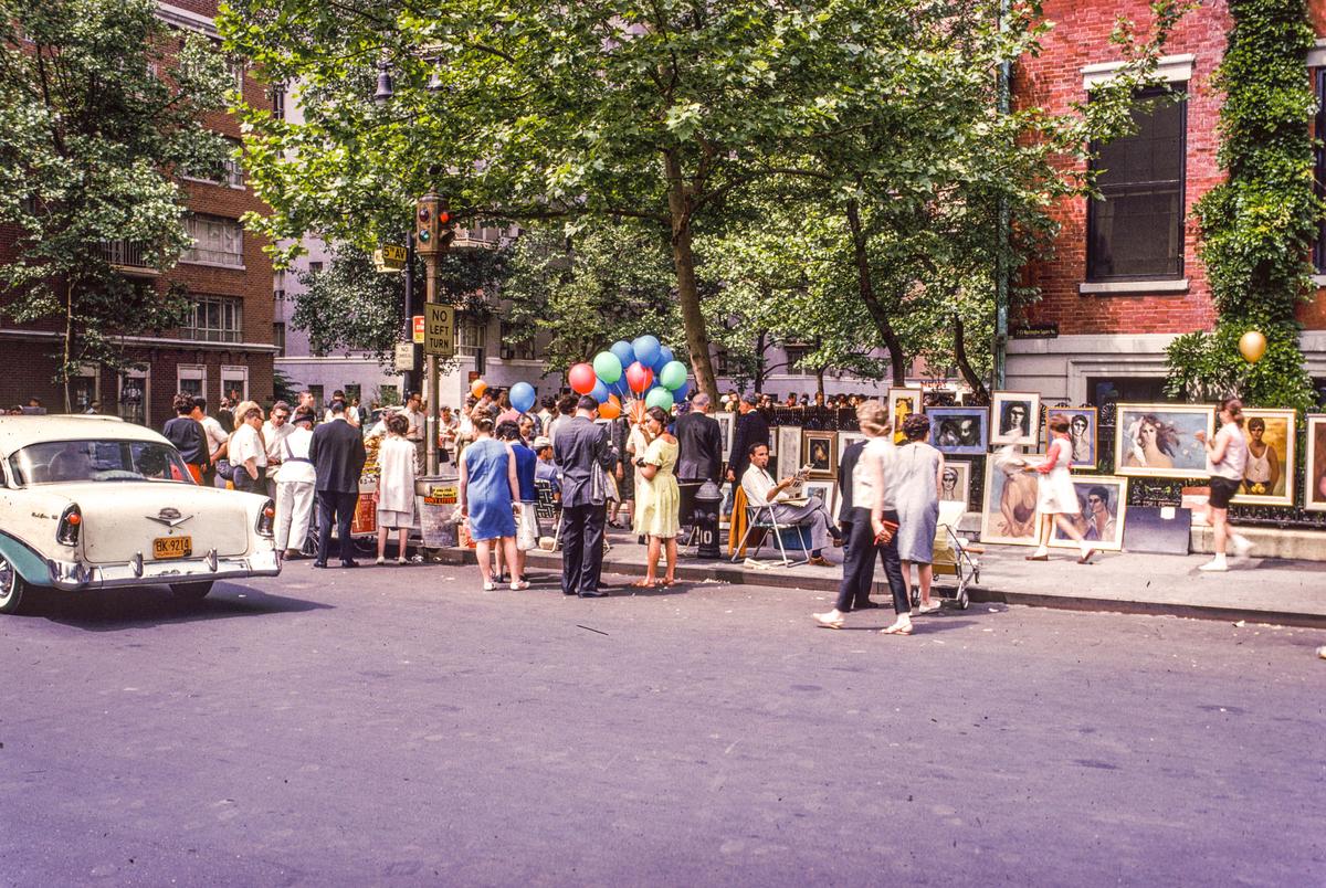 A street art fair is taking place on a sunny day with many people browsing paintings and a vintage car parked on the street.