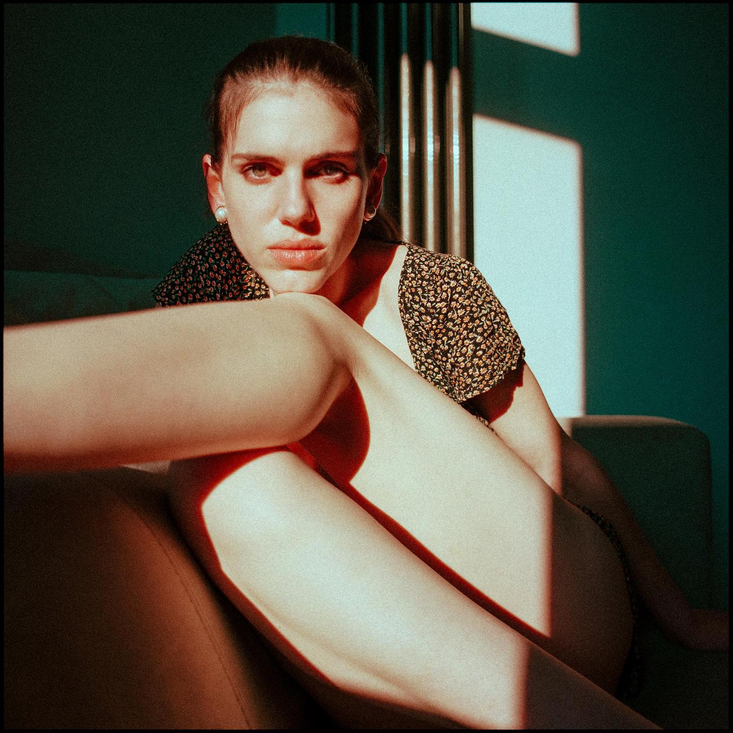 A woman sits on a couch with her legs pulled up to her chest, illuminated by harsh sunlight.