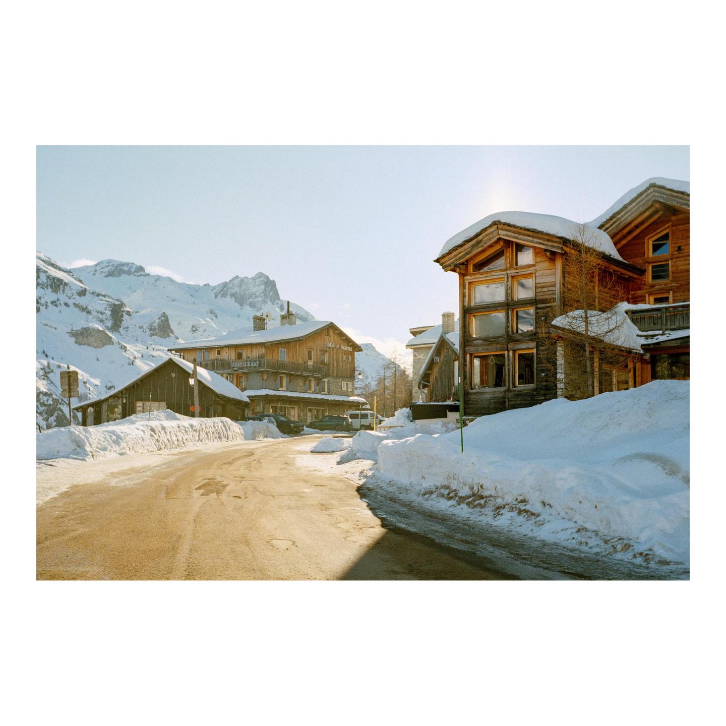 A snow-covered road leads past wooden buildings with mountains in the background.
