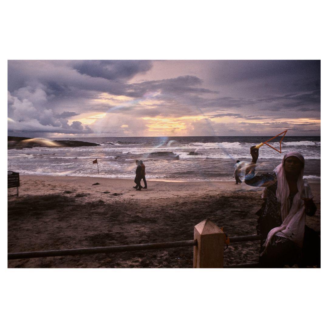 A woman holds a bubble wand on a beach with people walking near the ocean under a cloudy sky at sunset.