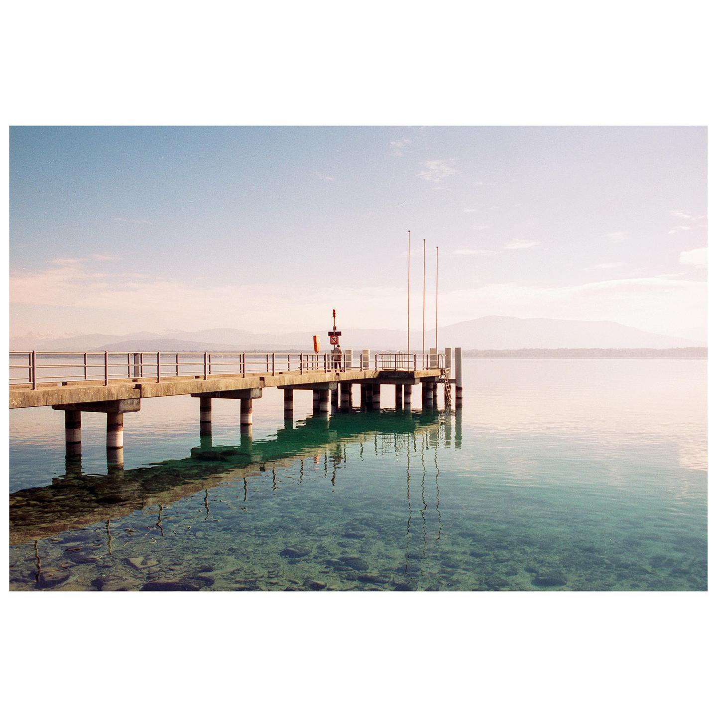 A concrete pier extends into clear, calm water with mountains in the distance under a hazy sky.