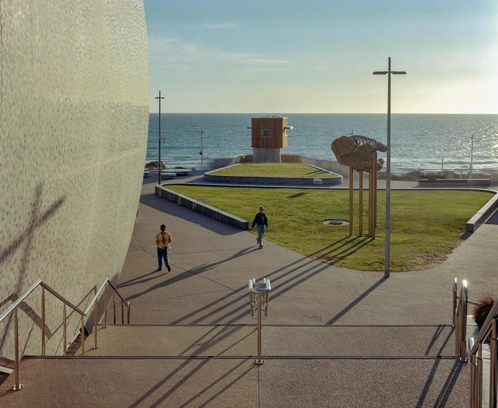 Two people walk on a paved path next to a grassy area and a large building, with the ocean visible in the background.