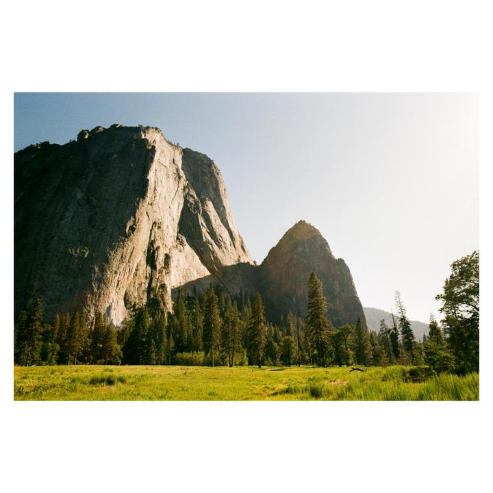 A large rock formation rises above a forest and a grassy field under a clear sky.