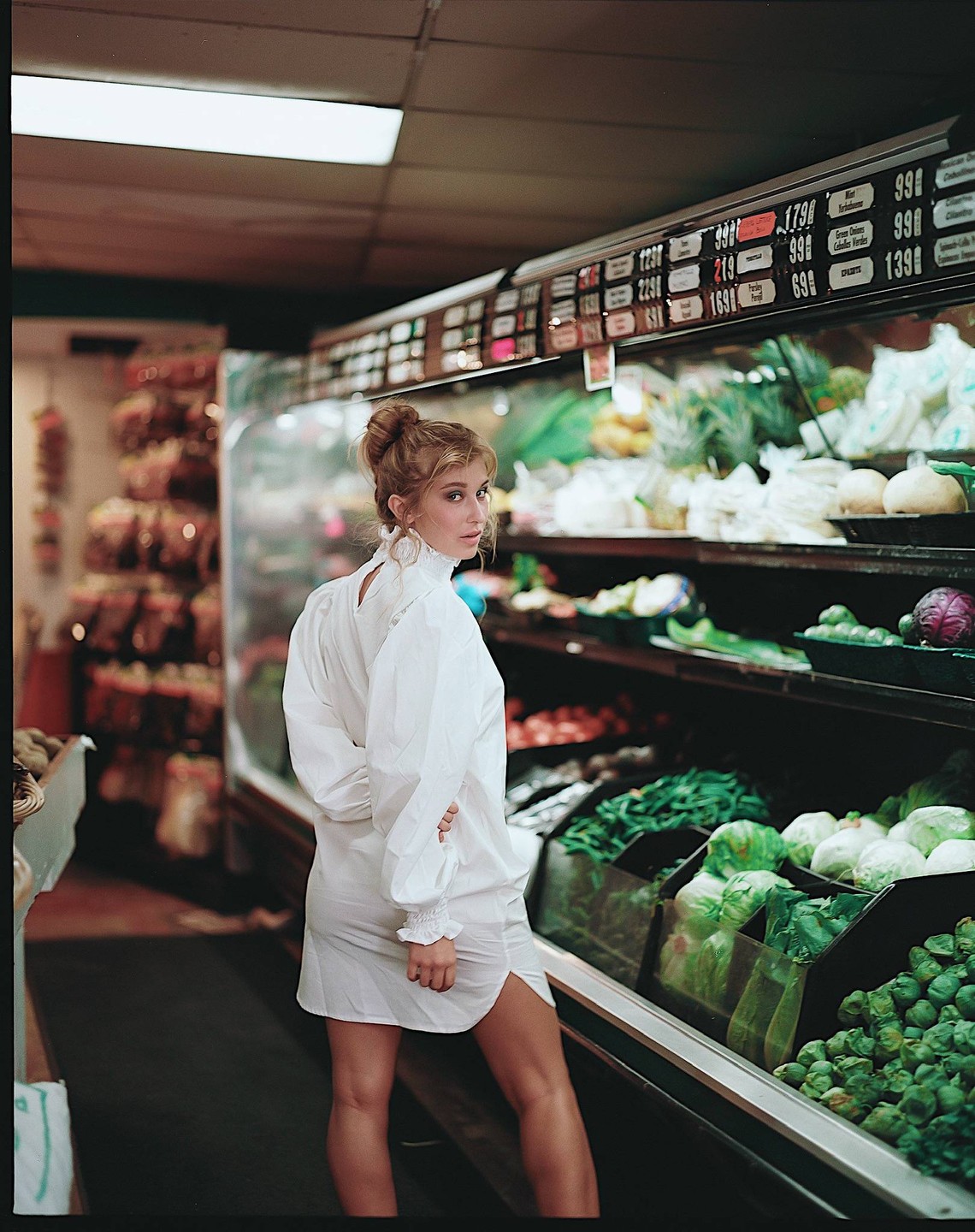 A woman in a white dress looks over her shoulder while standing in a grocery store aisle with produce.