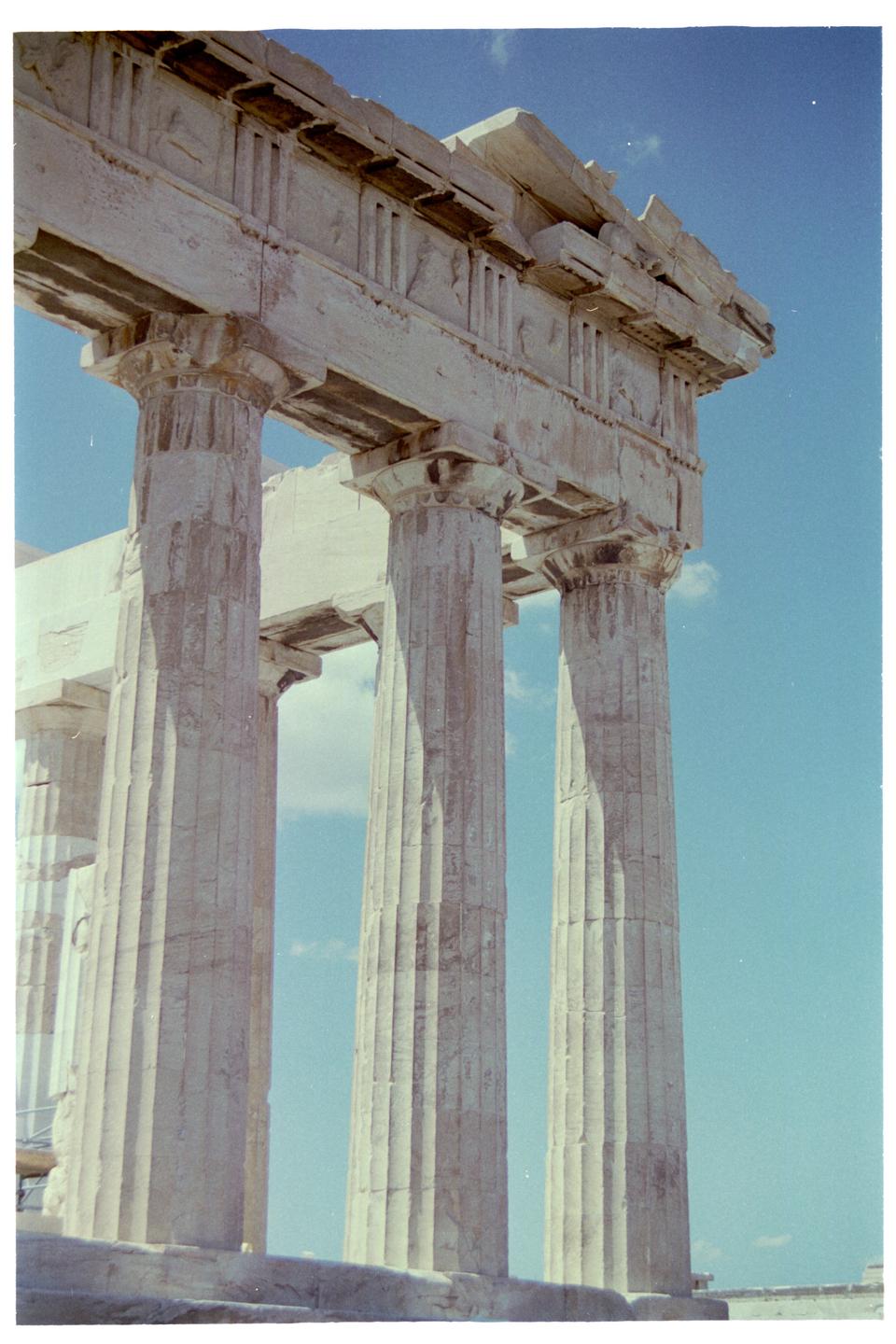 A low-angle view shows several large, fluted columns supporting a stone entablature against a clear blue sky.