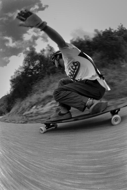 A person in a helmet and gloves is skateboarding downhill on a paved road with trees and clouds in the background.