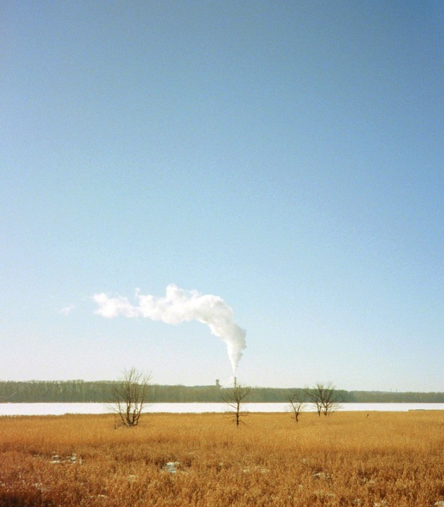 A plume of smoke rises from a distant factory chimney over a field of golden grass and a frozen lake.