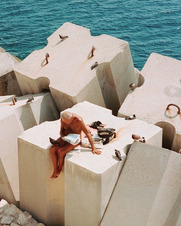 A man sits on concrete blocks by the sea, surrounded by pigeons.