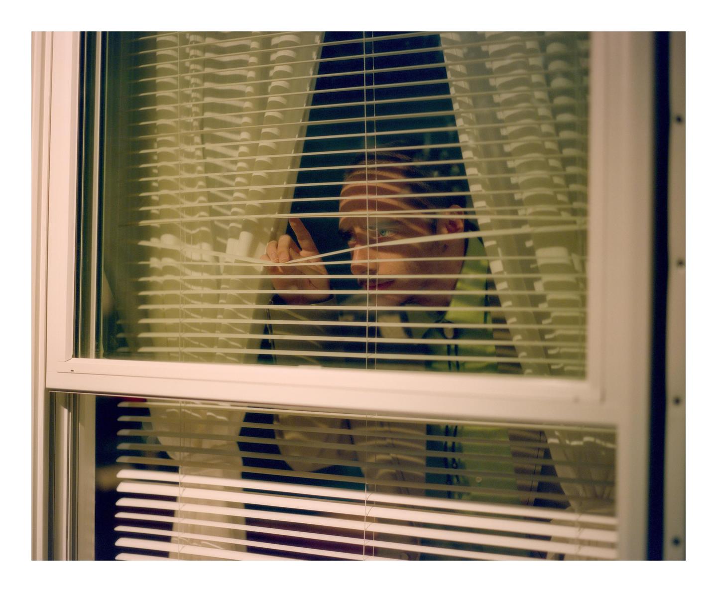 A man peeks through window blinds, with light stripes across his face.
