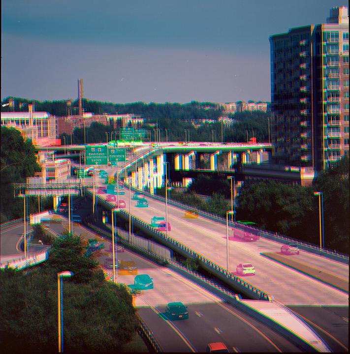 Cars drive on a multi-level highway system with buildings and trees in the background.