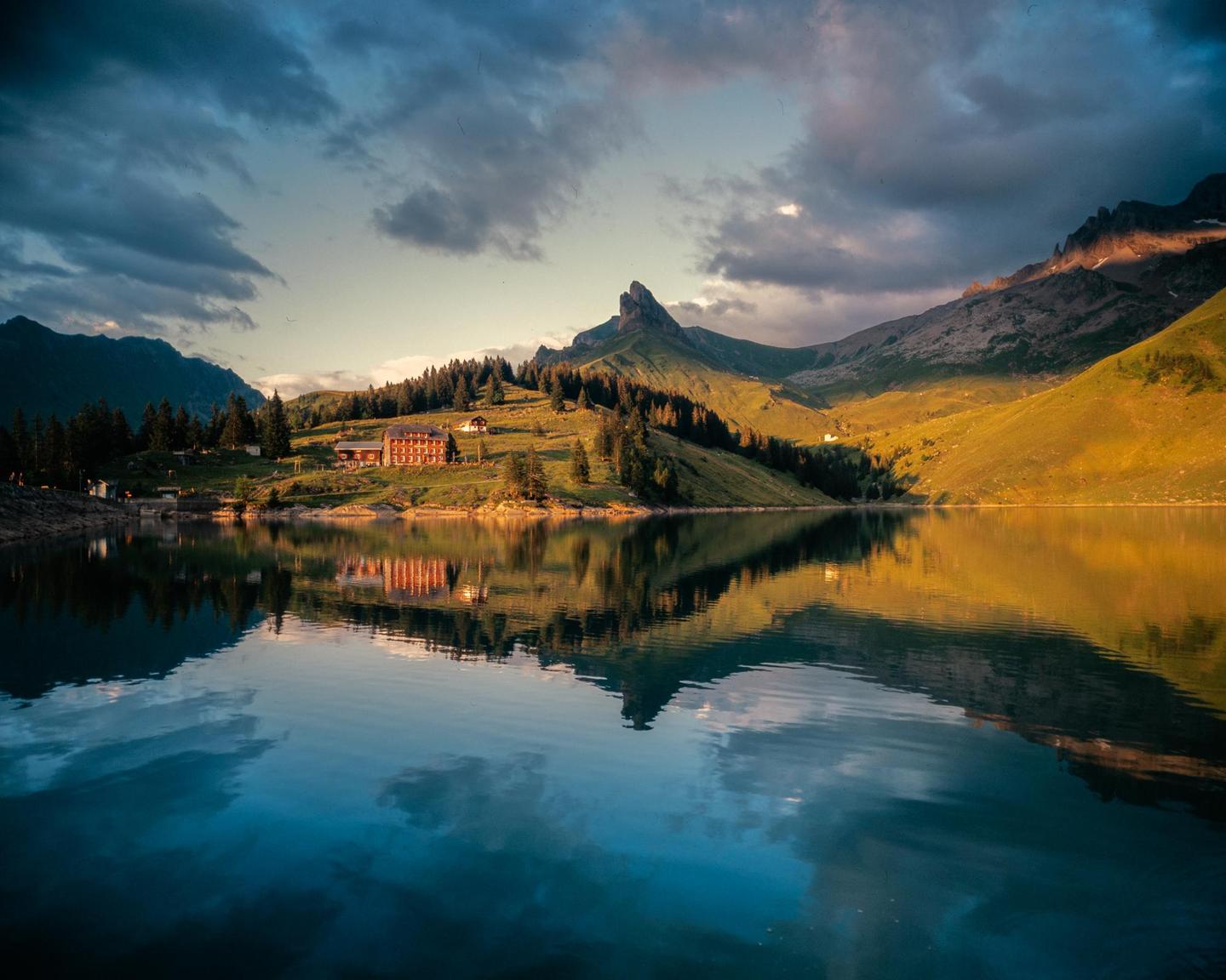 A large building with many windows sits on a grassy hill overlooking a lake, with mountains and clouds in the background.