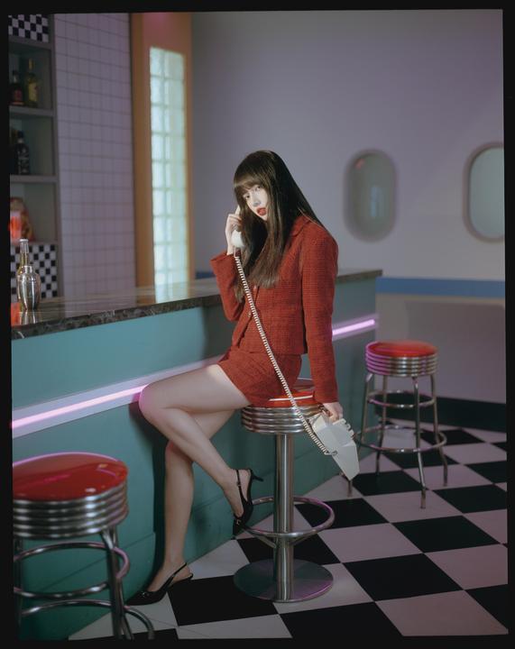 A woman in a red suit sits on a bar stool and talks on a white rotary phone in a diner.