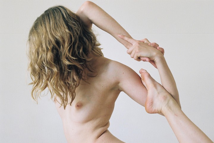 A woman with long wavy hair is shown from the back, holding her foot with her hands in a yoga pose.