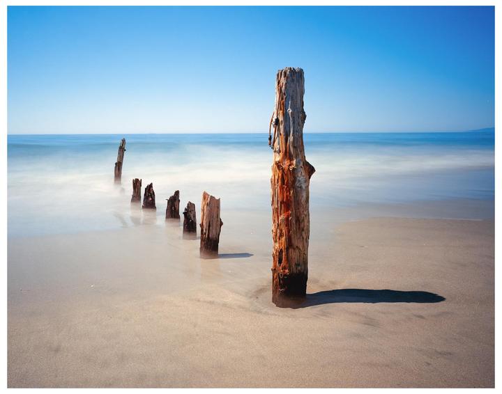 A line of wooden posts extends into the ocean from a sandy beach under a clear blue sky.