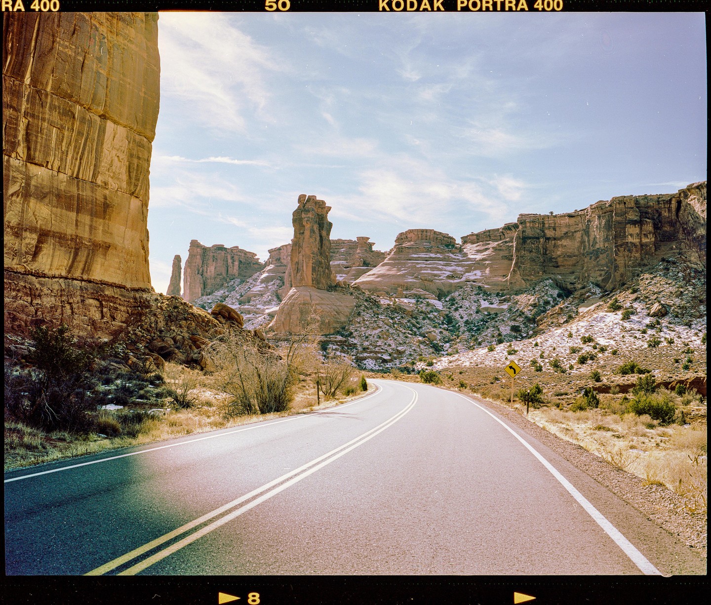 A winding road leads through a desert landscape with towering rock formations and patches of snow.