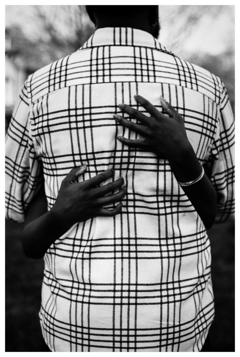 A person wearing a plaid shirt is embraced from behind by another person whose hands are visible.