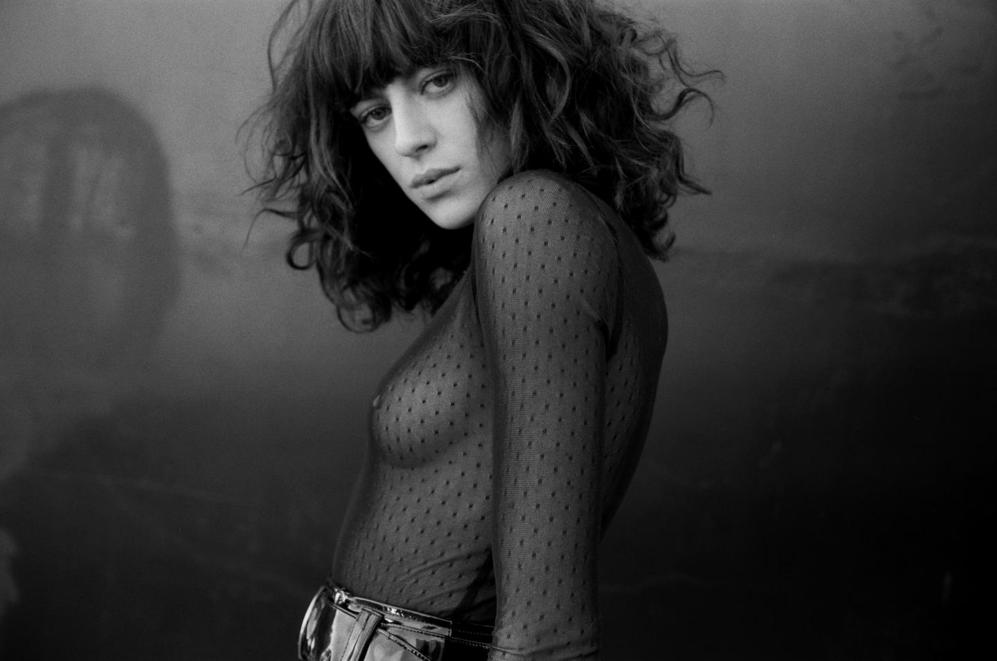 A woman with curly hair and bangs wears a sheer top and looks at the viewer.