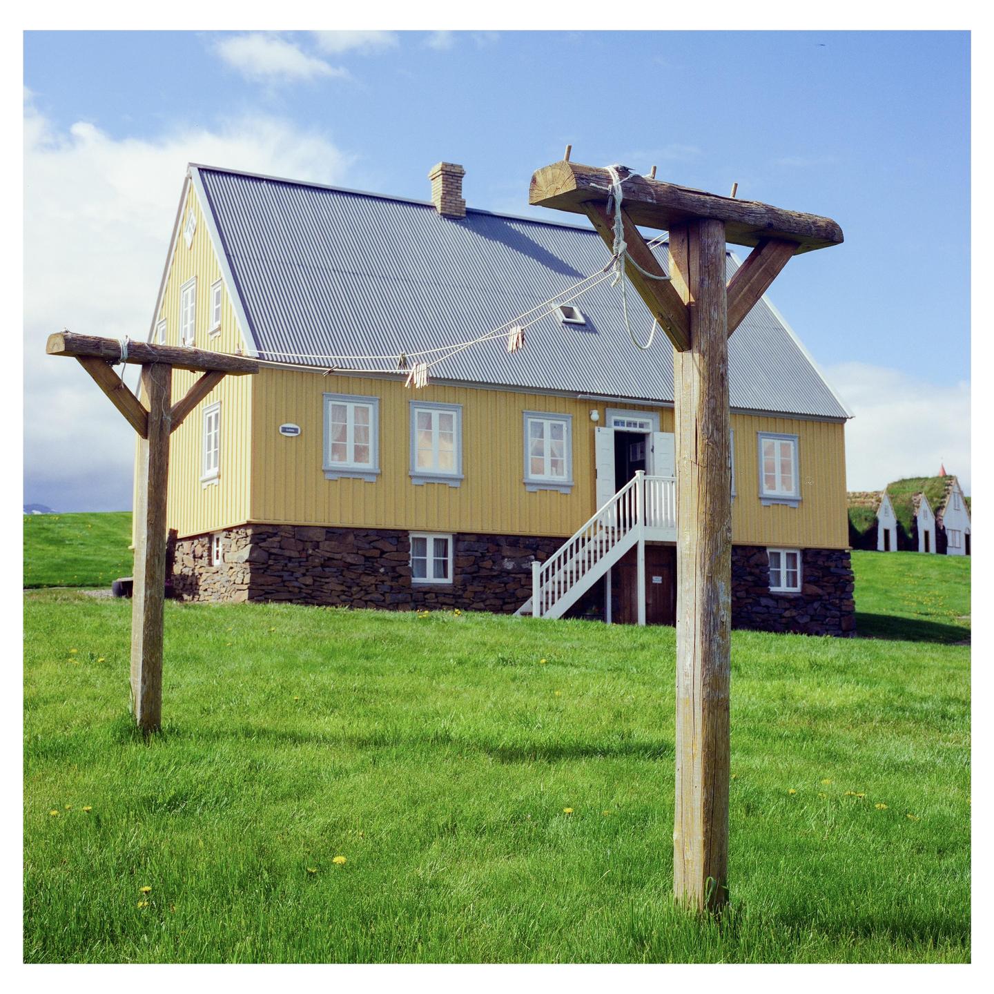 A yellow house with a gray roof stands on a green grassy hill under a blue sky.