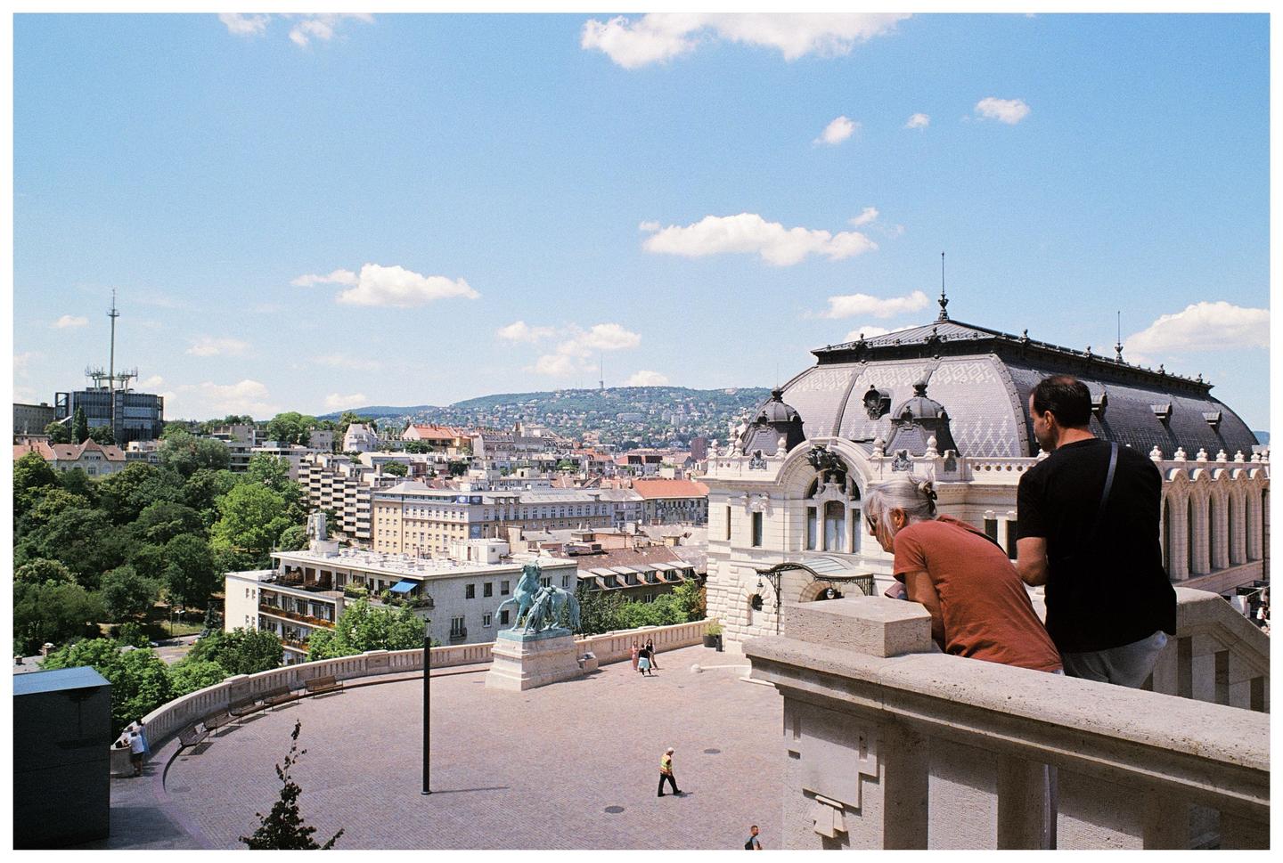 A man and a woman look out over a city from a balcony on a sunny day.