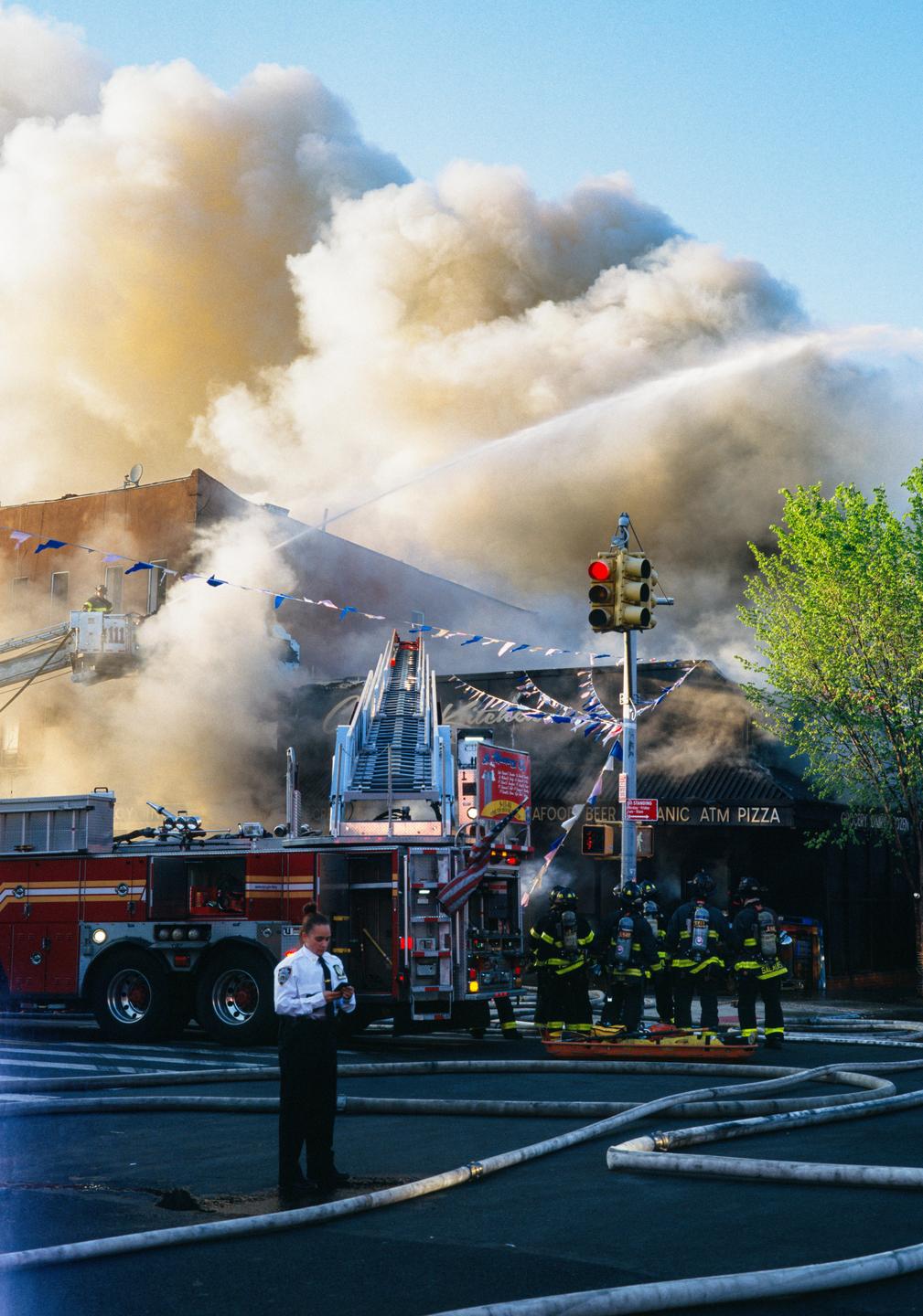 A fire truck is parked on a street with firefighters and a woman in uniform, while smoke billows from a building and water is sprayed.