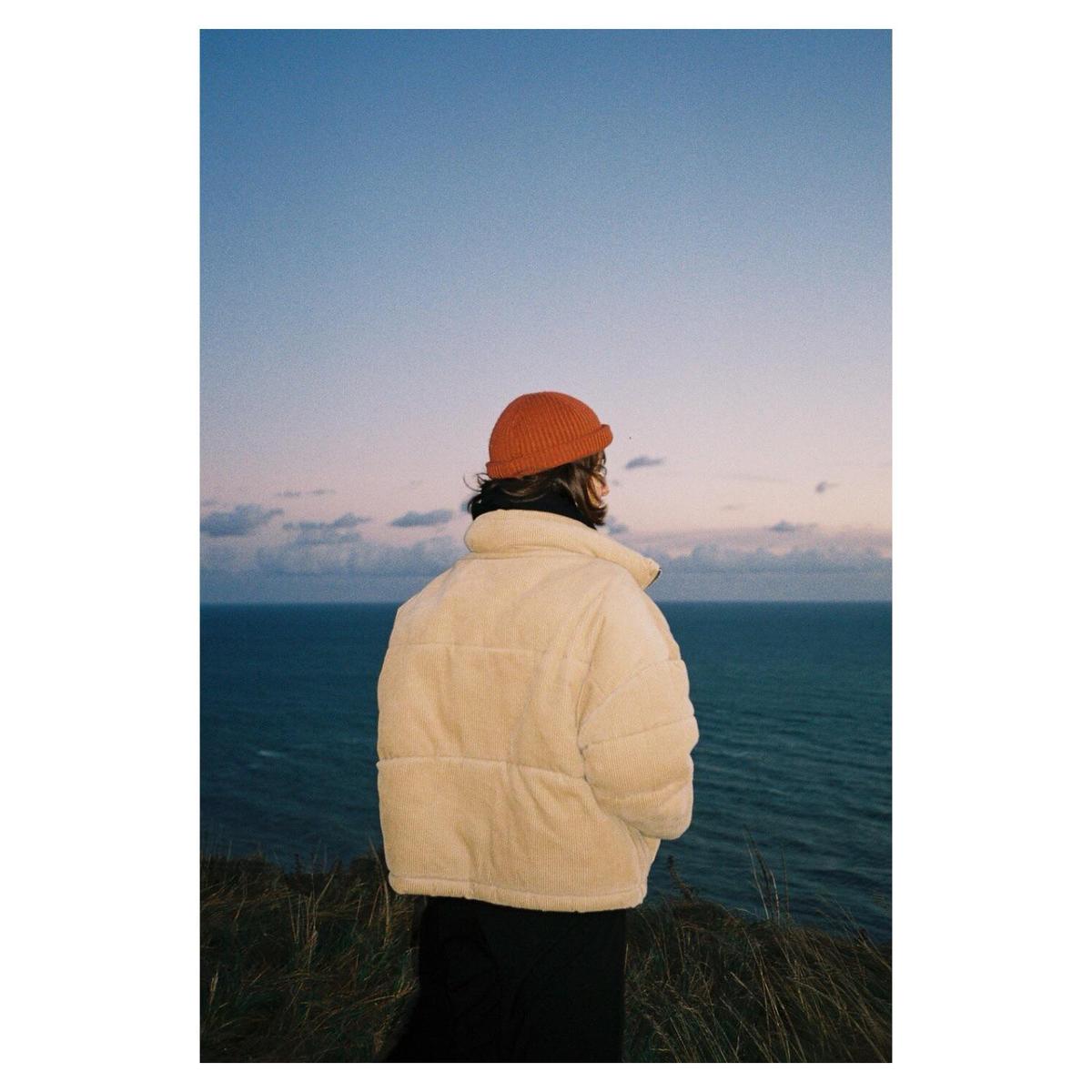 A person in a puffer jacket and beanie stands on a cliff overlooking the sea at dusk.