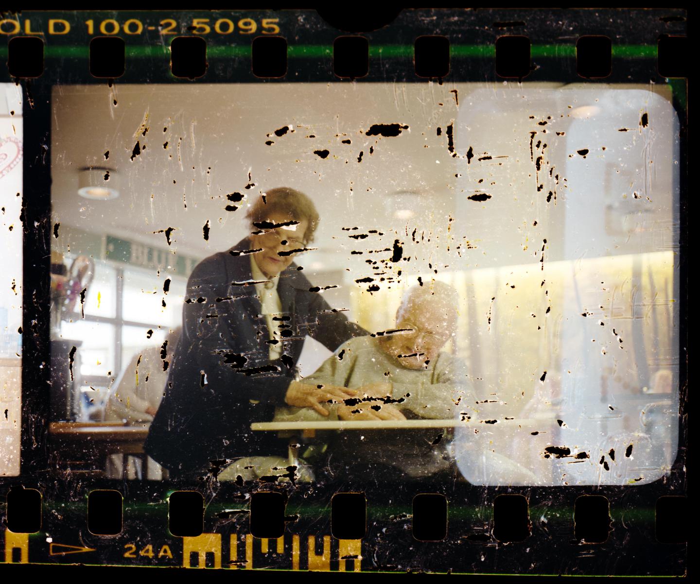 A woman helps an elderly man seated at a table.