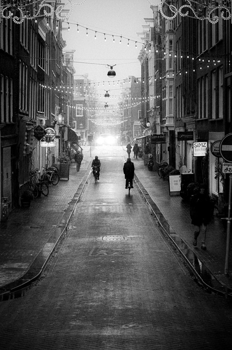 A street in Amsterdam is wet from rain, with string lights crisscrossing overhead and people walking and cycling.