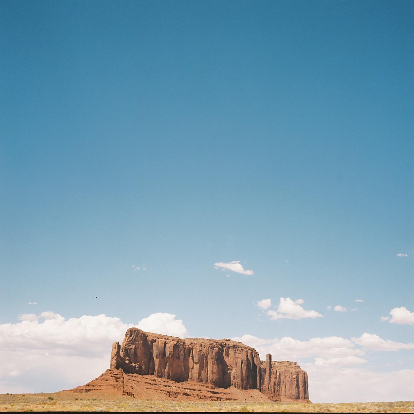 A large mesa rises from a flat desert landscape under a clear blue sky with scattered clouds.