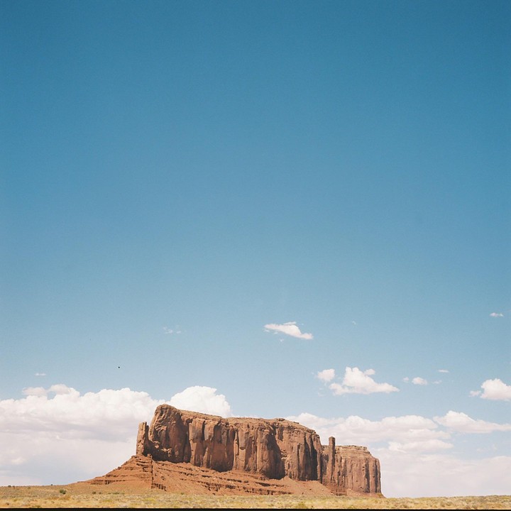 A large mesa rises from a flat desert landscape under a clear blue sky with scattered clouds.