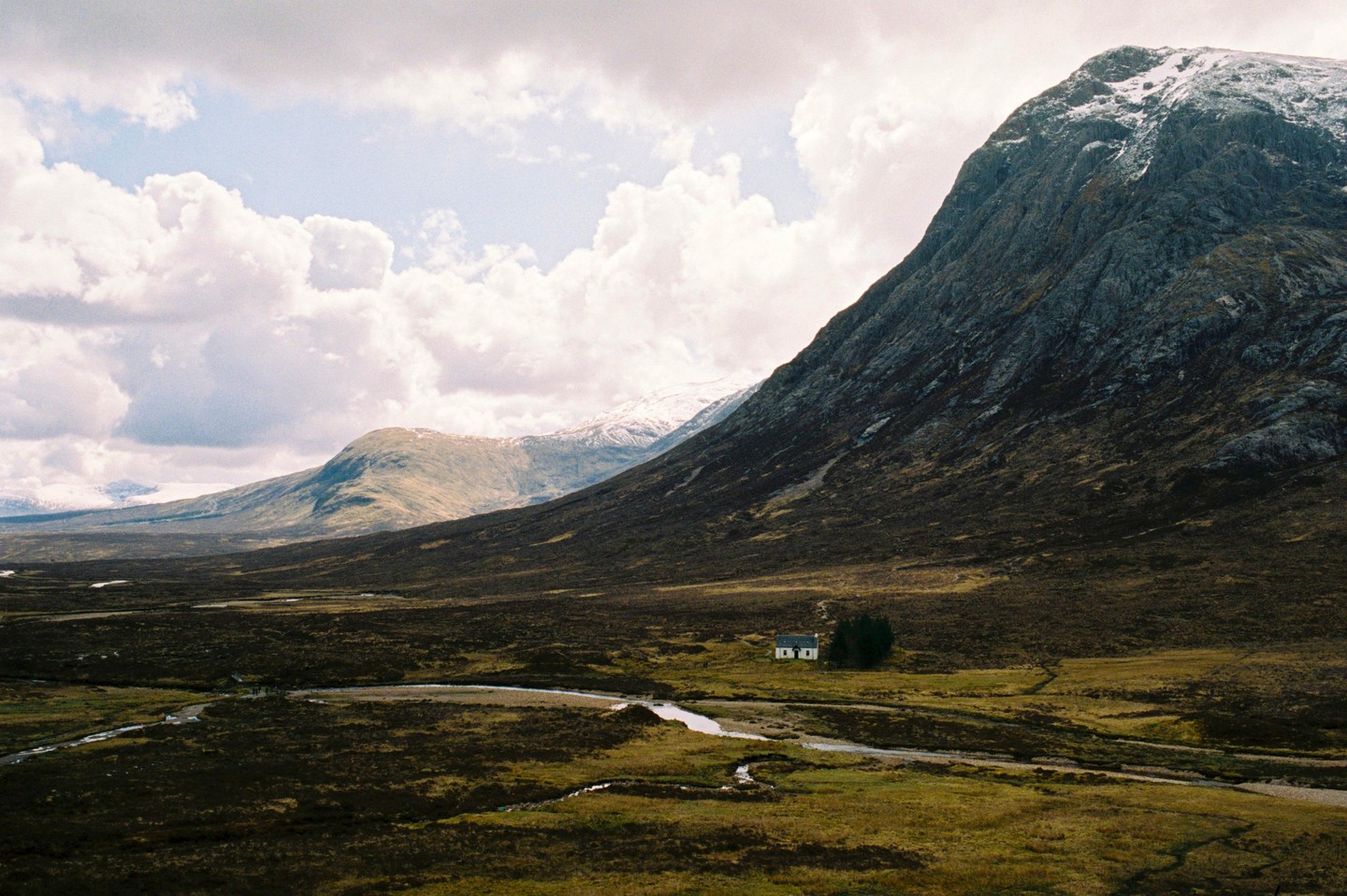 A small white house sits in a vast valley with a winding river, surrounded by mountains under a cloudy sky.