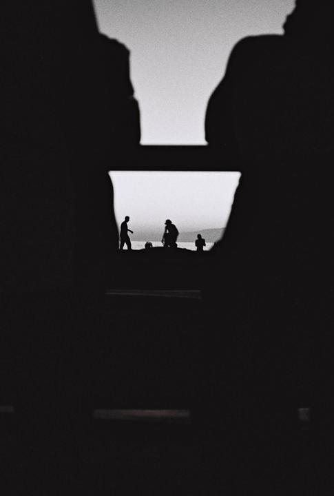 Three people are silhouetted against the ocean and sky, framed by dark shapes in the foreground.