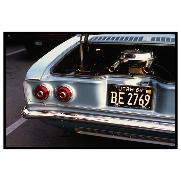 The rear of a light blue vintage car with its trunk open to reveal the engine is parked on asphalt.