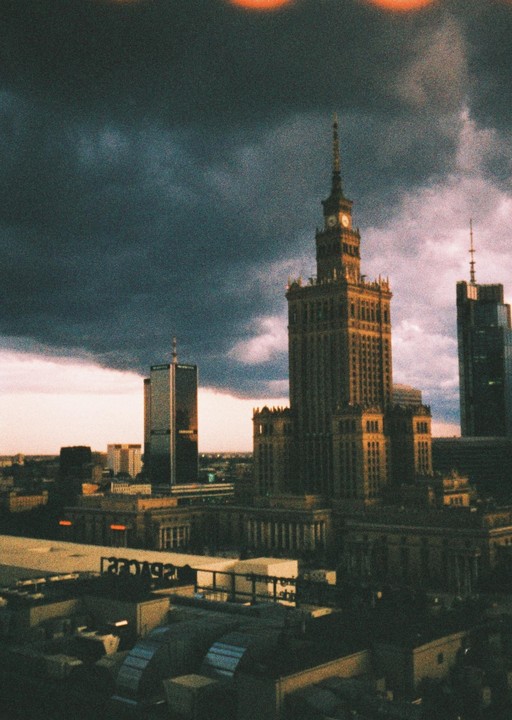 A tall, ornate building with a clock tower stands prominently against a dramatic, cloudy sky at dusk, surrounded by other city buildings.