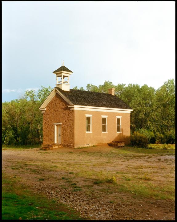 A small, old, adobe building with a bell tower stands in a dirt field surrounded by trees.