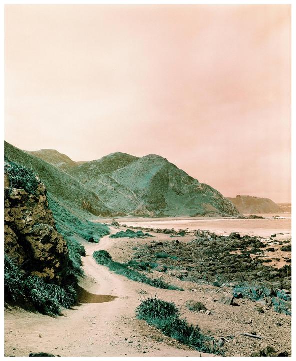 A dirt path winds along a rocky coastline with green hills under a pink sky.