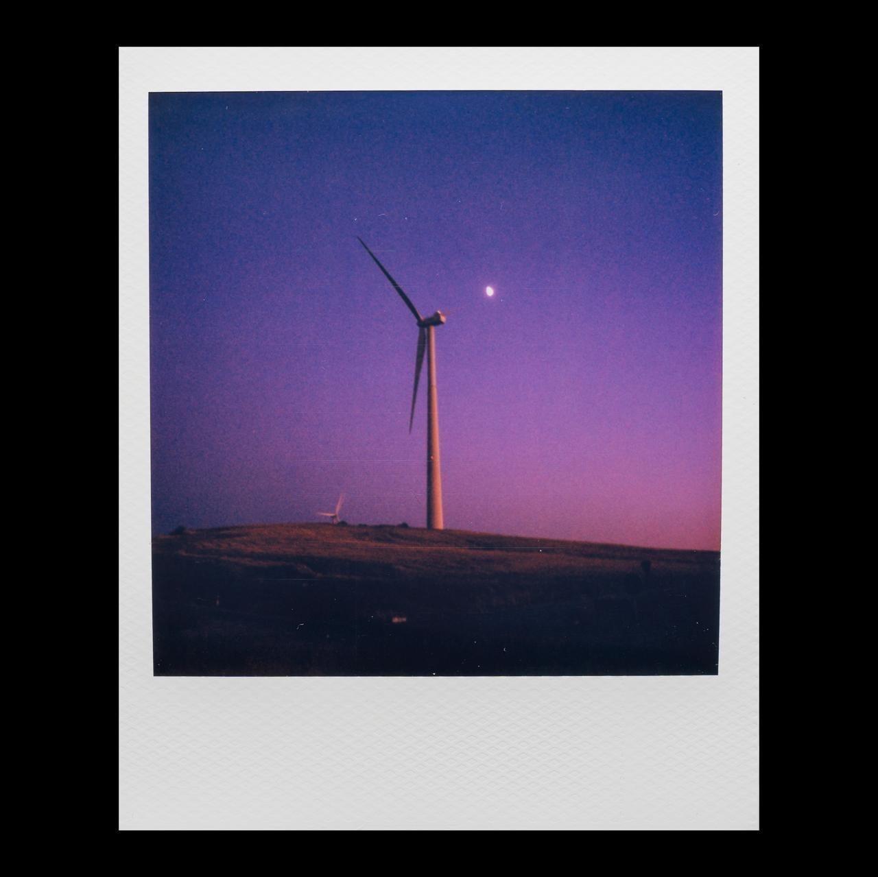A wind turbine stands on a hill under a purple and blue sky with a crescent moon.