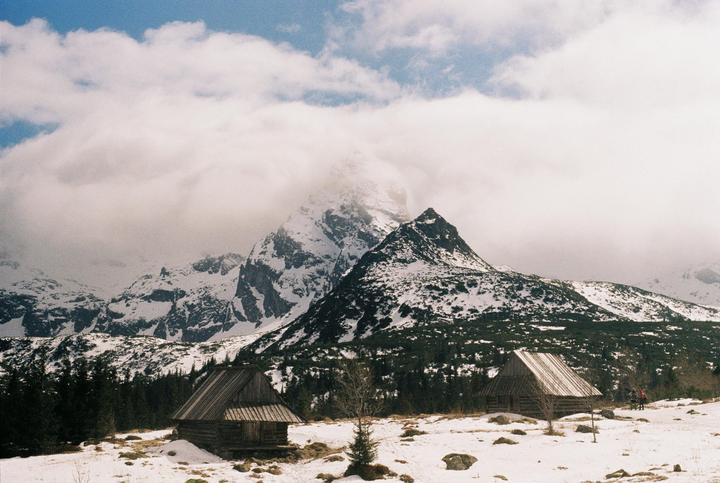 Two wooden cabins sit in a snowy landscape with mountains and clouds in the background.