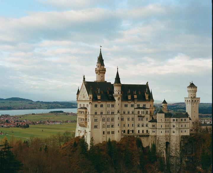 A large castle with multiple towers and spires stands on a rocky outcrop overlooking a valley with a lake and a village.